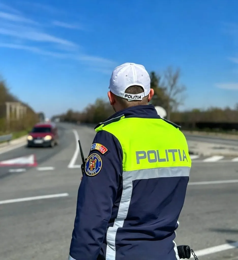 Un polițist cu uniformă albastră și vestă reflectorizantă galbenă stă pe marginea unei drumuri cu mașini în fundal sub un cer senin.