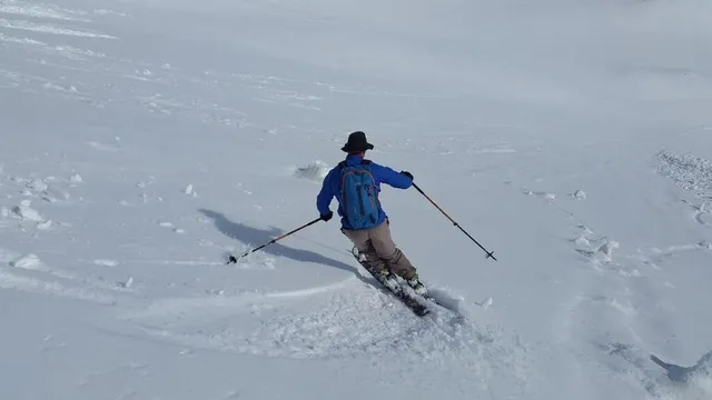 Un om cu sac de spate albastru și pantaloni bej alunecă pe zăpadă cu schiurile și bețele de schi.