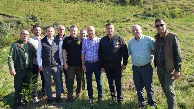 Un grup de nouă bărbați stă în picioare pe un teren verde în fața unui deal împădurit.