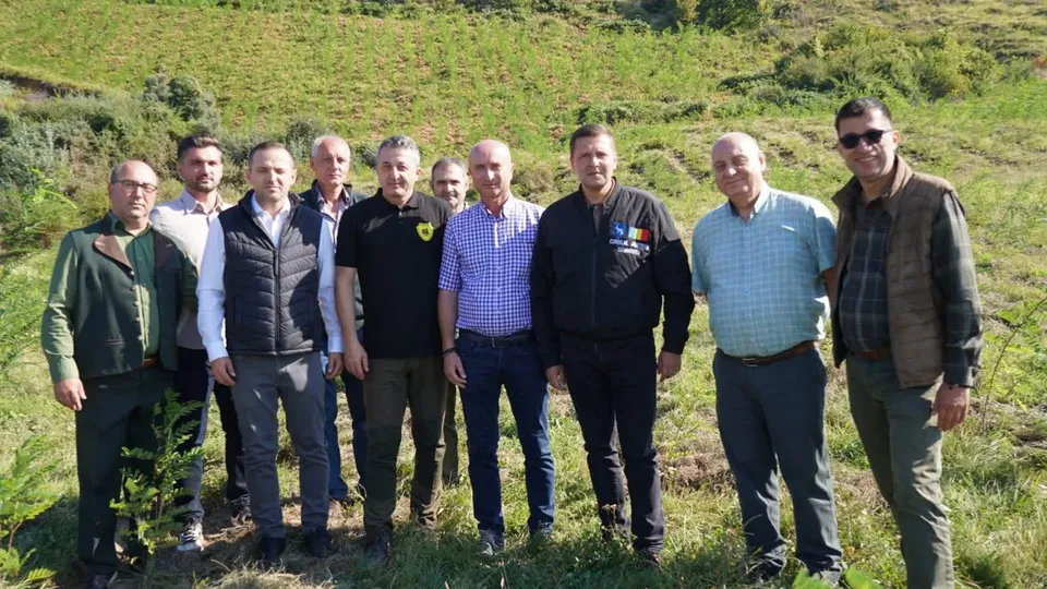 Un grup de nouă bărbați stă în picioare pe un teren verde în fața unui deal împădurit.