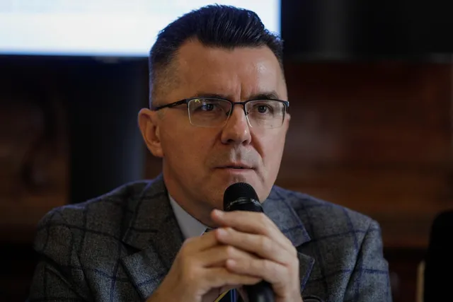 Un bărbat cu ochelari și sacou în croit gri vorbește într-un microfon ținut cu ambele mâini.