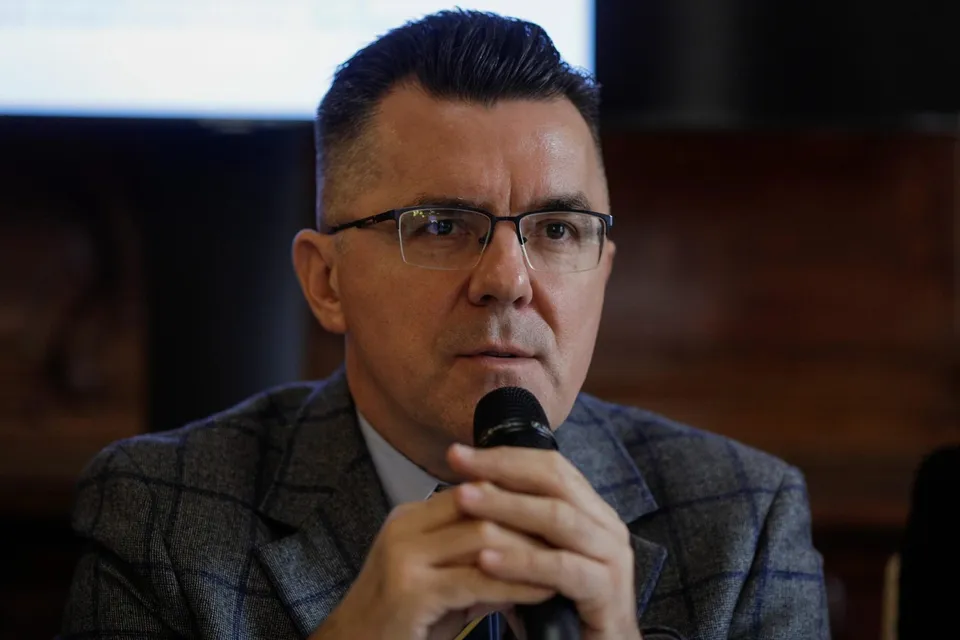 Un bărbat cu ochelari și sacou în croit gri vorbește într-un microfon ținut cu ambele mâini.