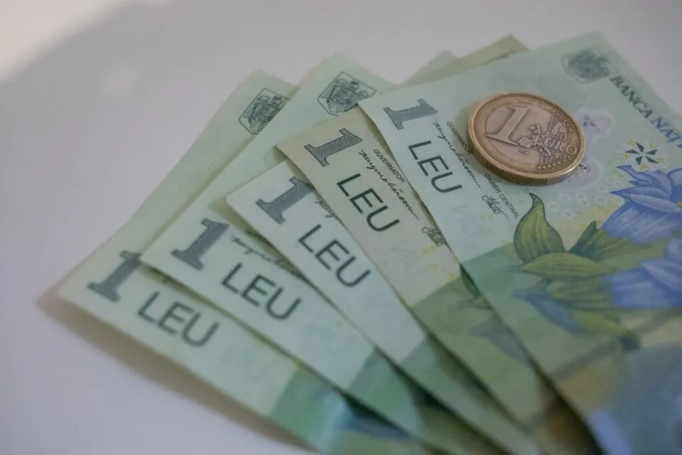 Mai multe bancnote de un leu românesc sunt întinse pe o suprafață albă lângă o monedă de un euro.