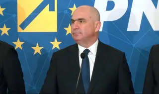 Un bărbat cu părul scurt și pielea deschisă poartă un costum negru, cămașă albă și cravată albastră în fața unui fundal albastru cu stele galbene.