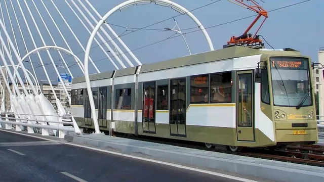 Un tramvai de culoare verde și alb circulă pe șine pe un pod cu structuri metalice albe curbe sub un cer senin.