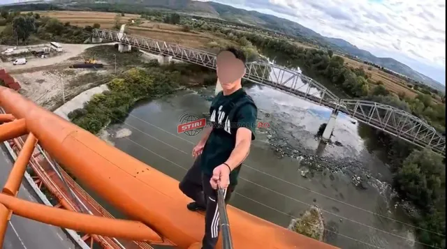 Un bărbat cu tricou închis și pantaloni închiși stă pe o structură metalică portocalie deasupra unui râu și a unui pod metalic.