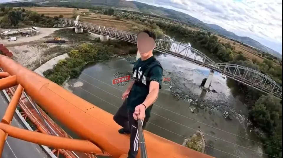 Un bărbat cu tricou închis și pantaloni închiși stă pe o structură metalică portocalie deasupra unui râu și a unui pod metalic.