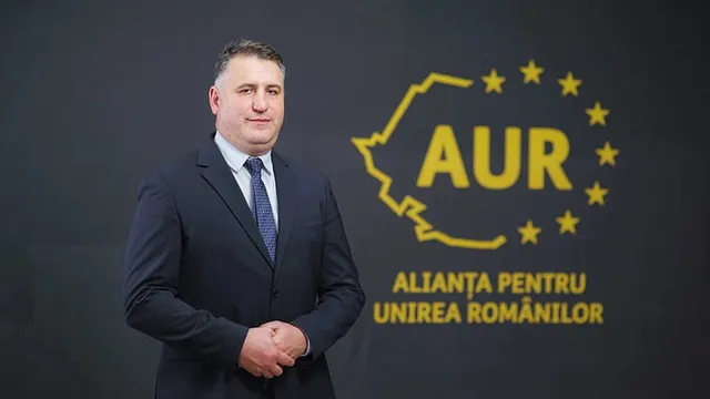 Un bărbat într-un costum închis la culoare și cravată stă în fața unui fundal negru cu un logo galben și textul Alianța pentru Unirea Românilor.