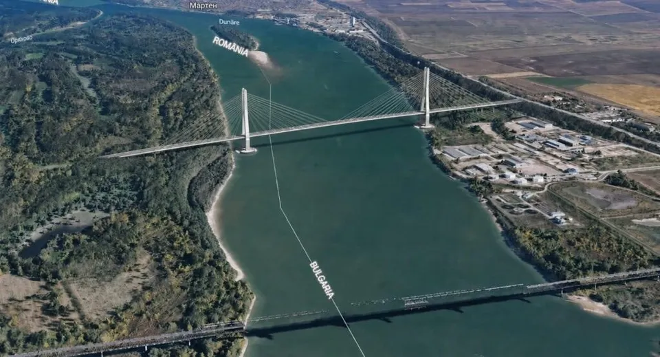 O vedere aeriană a unui fluviu lat traversat de un pod suspendat alb și un pod de tip viaduct, flancat de vegetație și zone industriale.