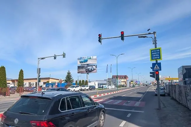 Un semafor suspendat de un stâlp metalic prezintă lumini roșii deasupra unei artere rutiere cu mașini și marcaje pe asfalt sub un cer senin.