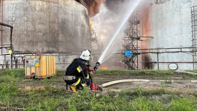 Un pompier în echipament de protecție stă în poziție de genunchi și direcționează un jet de apă către un rezervor mare de depozitare.