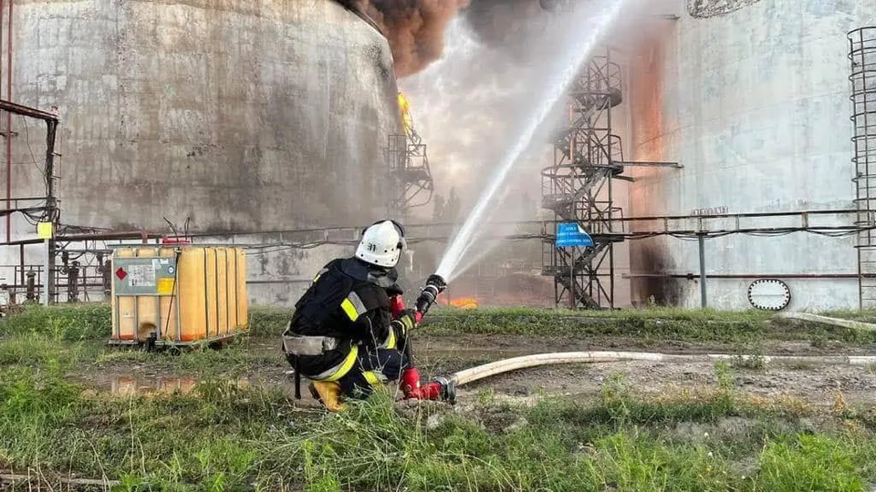 Un pompier în echipament de protecție stă în poziție de genunchi și direcționează un jet de apă către un rezervor mare de depozitare.
