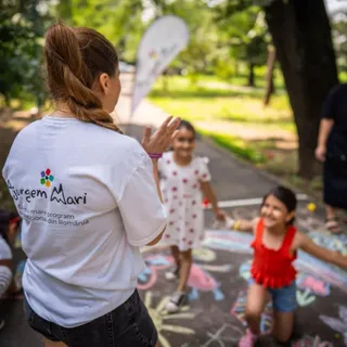 O femeie cu părul legat într-o coadă de cal poartă un tricou alb și ridică mâinile spre o fată într-o rochie albă cu puncte roșii.
