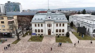 O clădire albă cu acoperiș gri este situată într-o piață pavată unde se află câțiva oameni și arbori fără frunze.