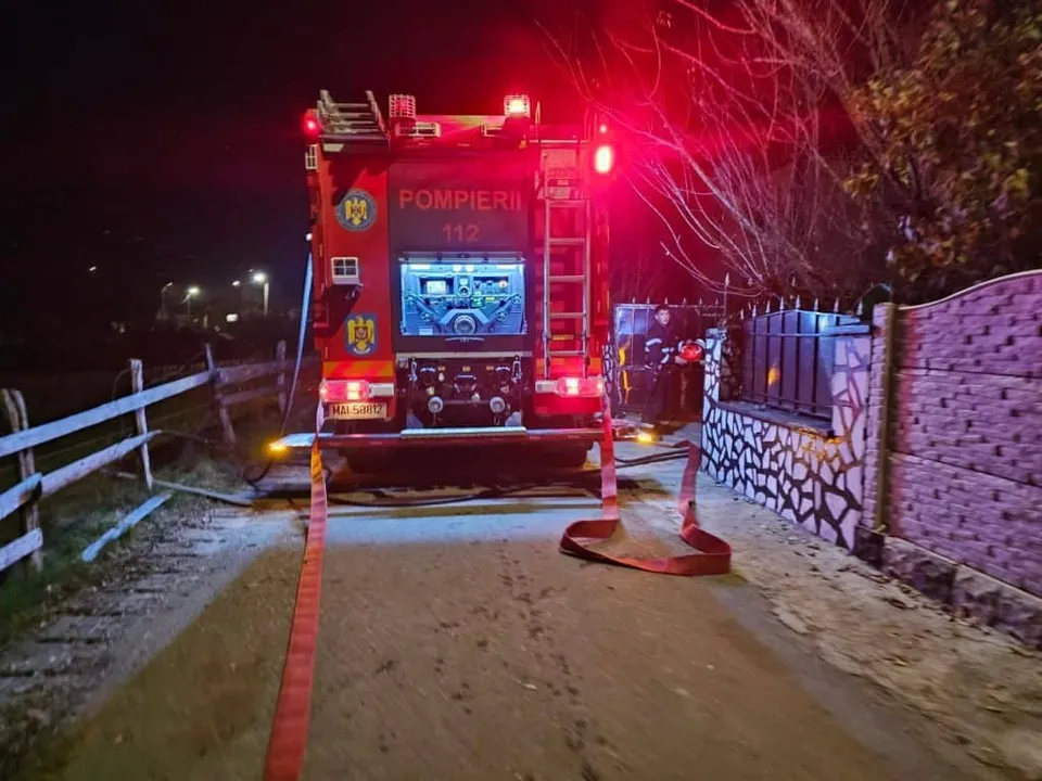 Un camion de pompieri cu luminile roșii aprinse stă pe un drum, cu furtunuri întinse pe sol lângă un gard.
