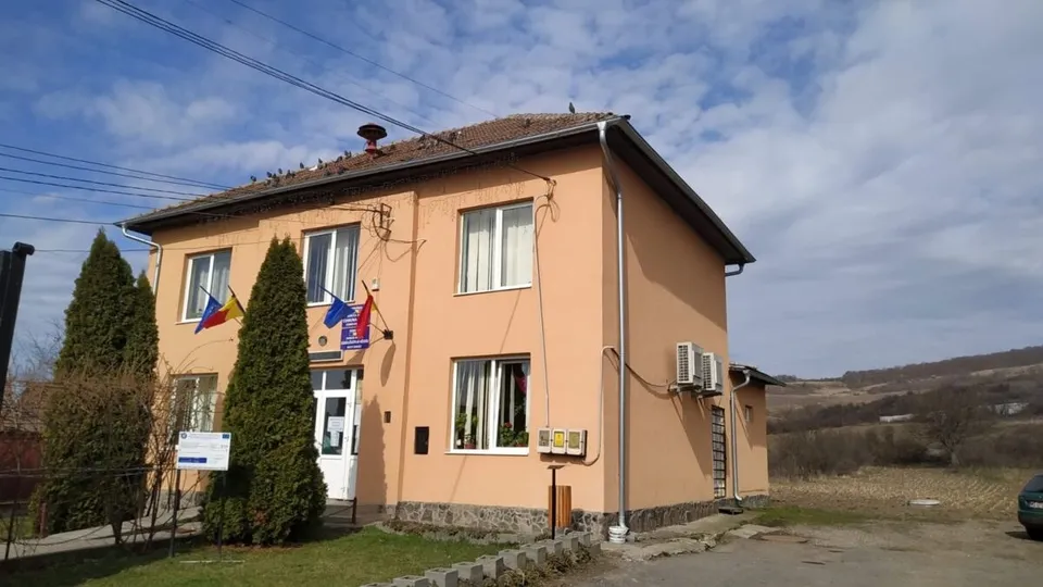 O clădire cu fațadă portocalie, având ferestre albe, două steaguri și două unități de aer condiționat montate pe perete, sub un cer înnorat.