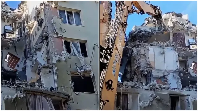 O excavatoare portocalie lucrează lângă un imobil de beton parțial demolat cu ferestre și pereți expuși.