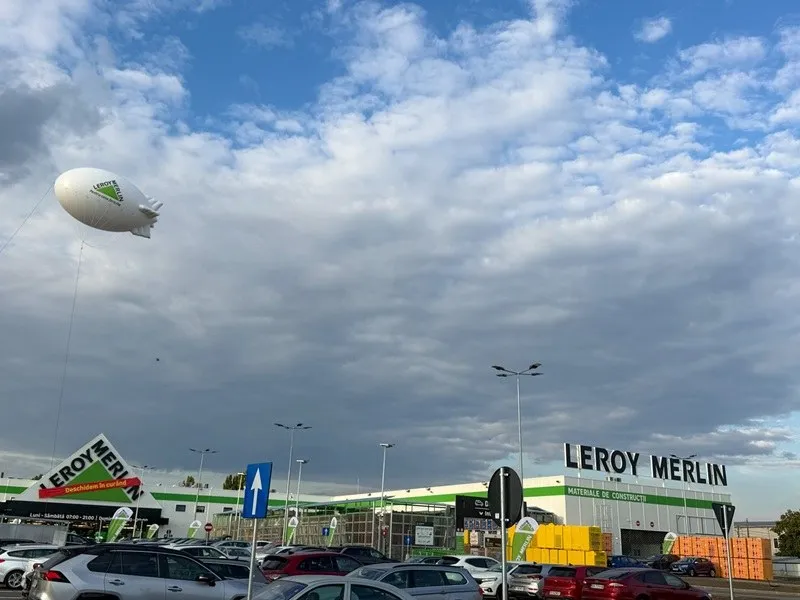 O imagine exterioară a unui magazin Leroy Merlin, cu o parcare plină de mașini și un dirijabil alb cu logo-ul companiei plutind deasupra.