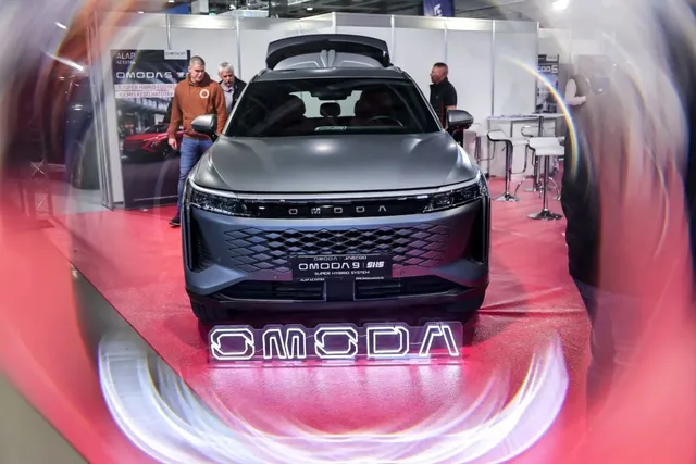 Un SUV gri este expus pe un covor roșu sub un logo luminos cu textul OMODA, în prezența unor persoane în fundal.