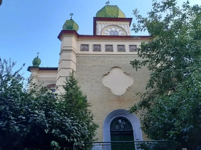 O clădire din cărămidă deschisă la culoare cu cupole verzi și un arc de intrare este parțial acoperită de vegetație verde.