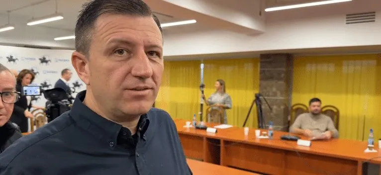 Un bărbat cu cămașă închisă la culoare privește spre cameră, în timp ce în fundal se văd persoane la mese într-o încăpere cu panouri galbene.