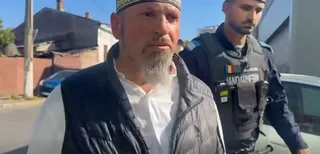 Un bărbat cu barbă și vestă neagră stă în prim-plan, în timp ce un oficial de poliție în uniformă gri stă în spatele lui.