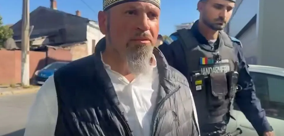 Un bărbat cu barbă și vestă neagră stă în prim-plan, în timp ce un oficial de poliție în uniformă gri stă în spatele lui.