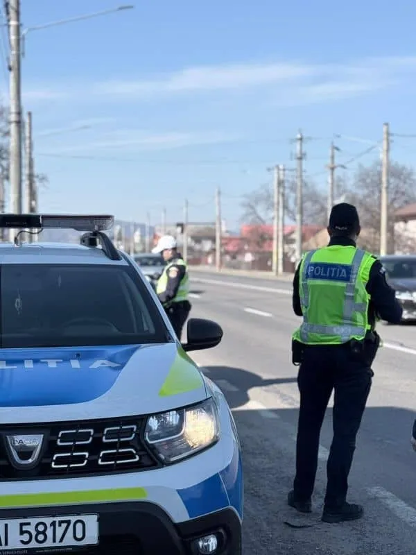 Un polițist cu vestă reflectorizantă verde stă pe marginea unui drum lângă o mașină de poliție albă și albastră sub un cer senin.