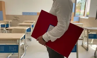 Un bărbat cu cămașă albă poartă un obiect dreptunghiular roșu prin interiorul unei săli de clasă cu bănci albe și albastre.
