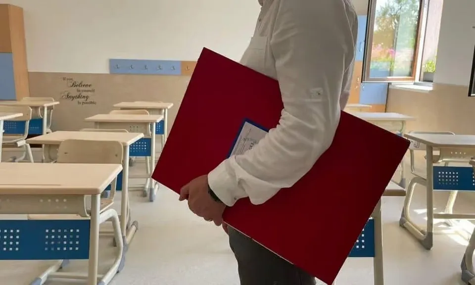 Un bărbat cu cămașă albă poartă un obiect dreptunghiular roșu prin interiorul unei săli de clasă cu bănci albe și albastre.