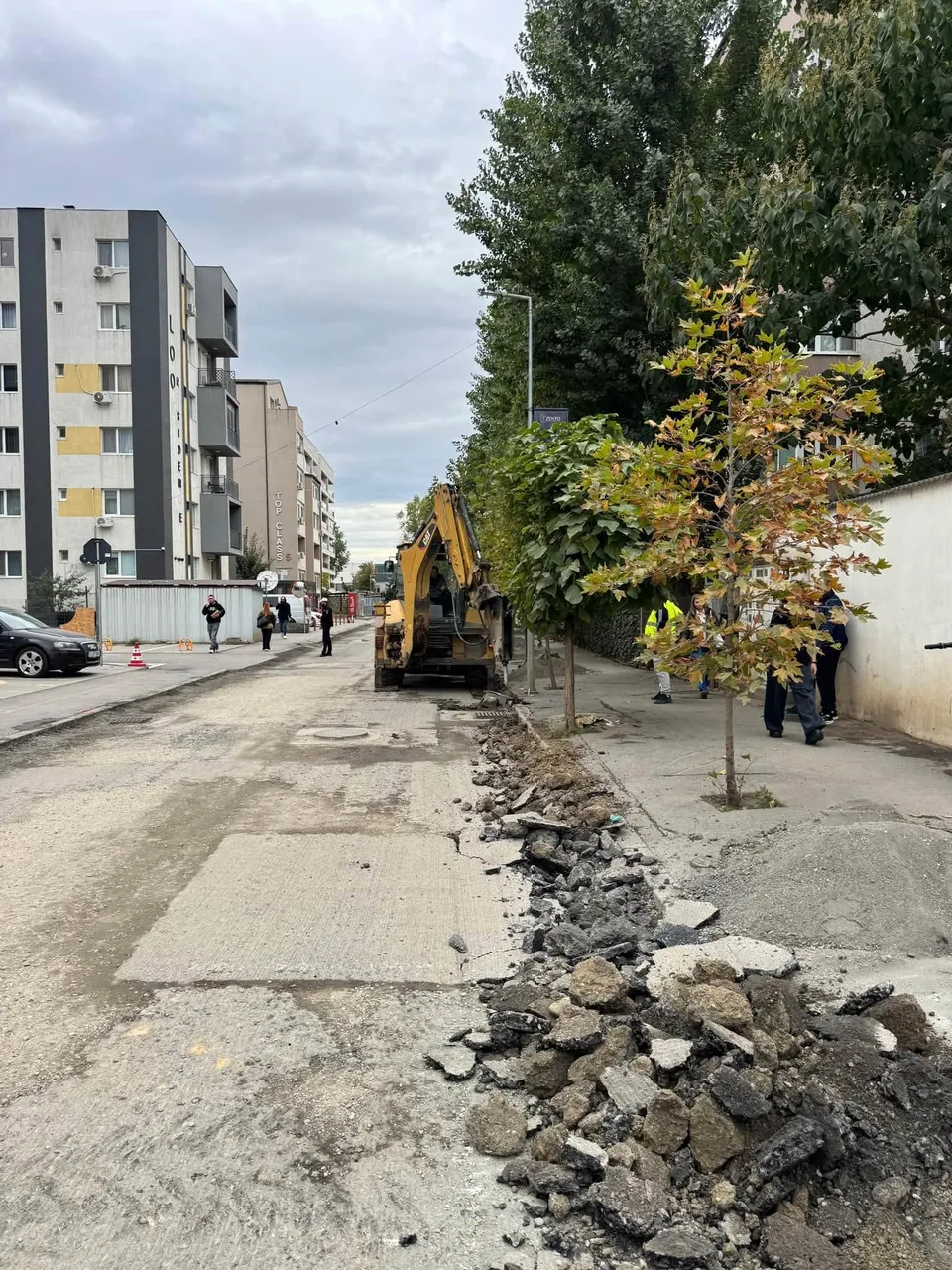 O excavatoare galbenă lucrează pe un drum de asfalt parțial demolat lângă un trotuar și clădiri de apartamente înalte sub un cer înnorat.