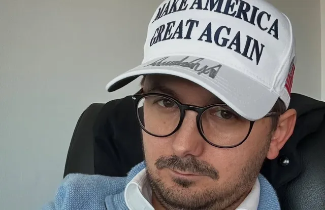 Un bărbat cu ochelari, mustășuș și pălărie albă cu inscripția 'MAKE AMERICA GREAT AGAIN' este pozitionat în fața unui fundal gri deschis.