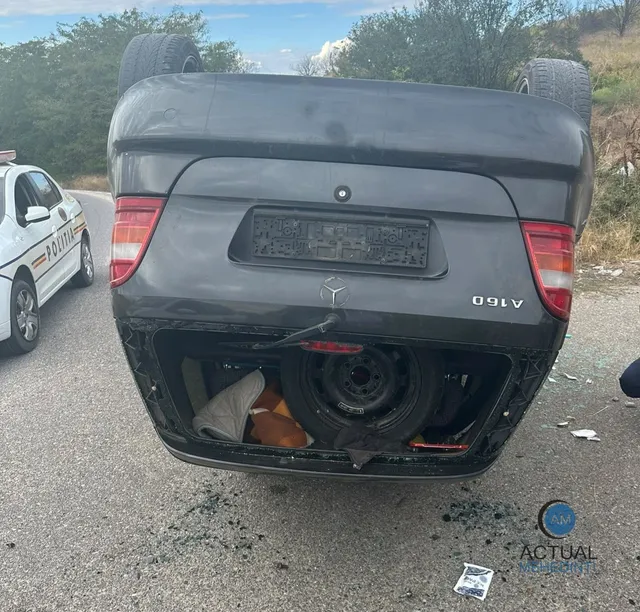 O mașină gri răsturnată pe șosea, cu portbagajul deschis, lângă o mașină de poliție albă.