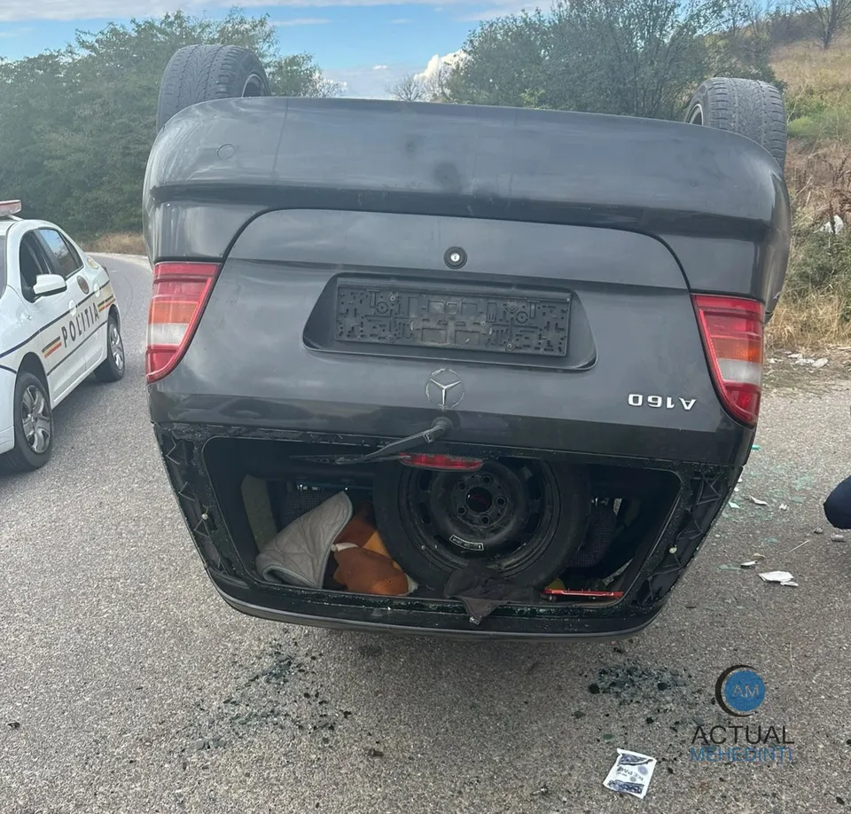 O mașină gri răsturnată pe șosea, cu portbagajul deschis, lângă o mașină de poliție albă.