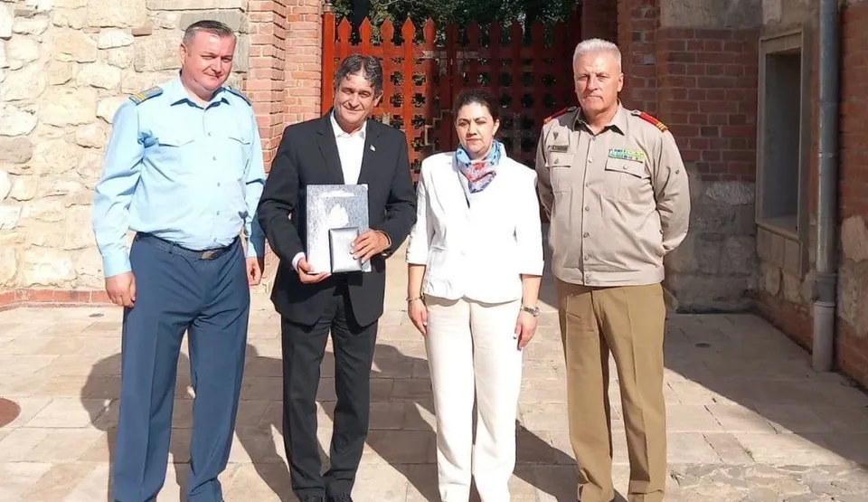 Patru persoane stau în rând pe o suprafață pavată în fața unei clădiri din piatră și cărămidă, un bărbat purtând un costum negru ține un cadou încadrat.