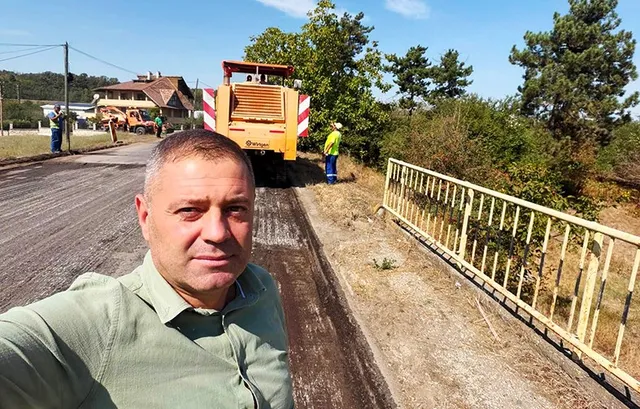Un bărbat în cămașă verde face un selfie pe un drum proaspăt asfaltat, lângă un utilaj de construcții și vegetație abundentă sub cerul senin.