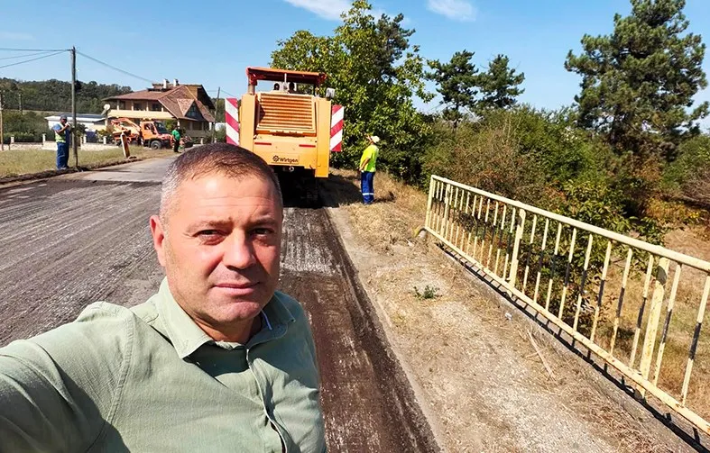 Un bărbat în cămașă verde face un selfie pe un drum proaspăt asfaltat, lângă un utilaj de construcții și vegetație abundentă sub cerul senin.
