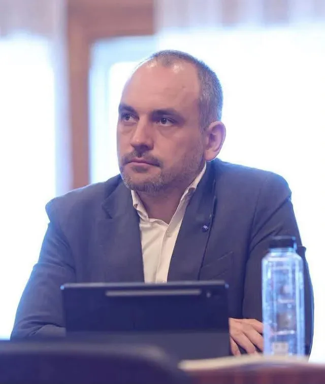 Un bărbat cu barbă scurtă, purtând sacou închis și cămașă albă, stă la o masă lângă o sticlă de apă și un dispozitiv electronic negru.
