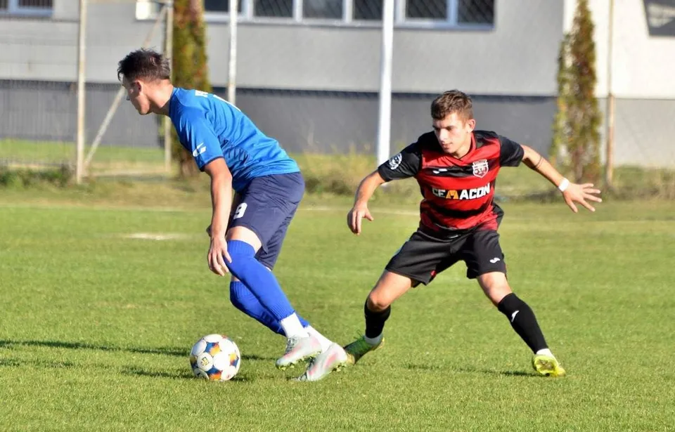 Un jucător în echipament albastru conduce mingea pe un teren de fotbal, fiind urmărit de un adversar îmbrăcat în echipament negru și roșu.