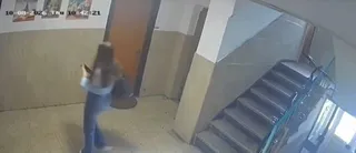 O persoană cu părul lung, purtând bluză deschisă la culoare și pantaloni albaștri, merge pe un hol lângă o scară interioară.