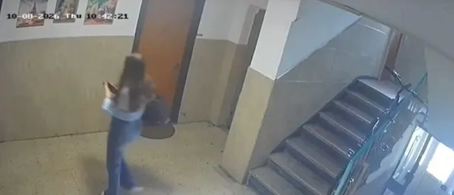 O persoană cu părul lung, purtând bluză deschisă la culoare și pantaloni albaștri, merge pe un hol lângă o scară interioară.