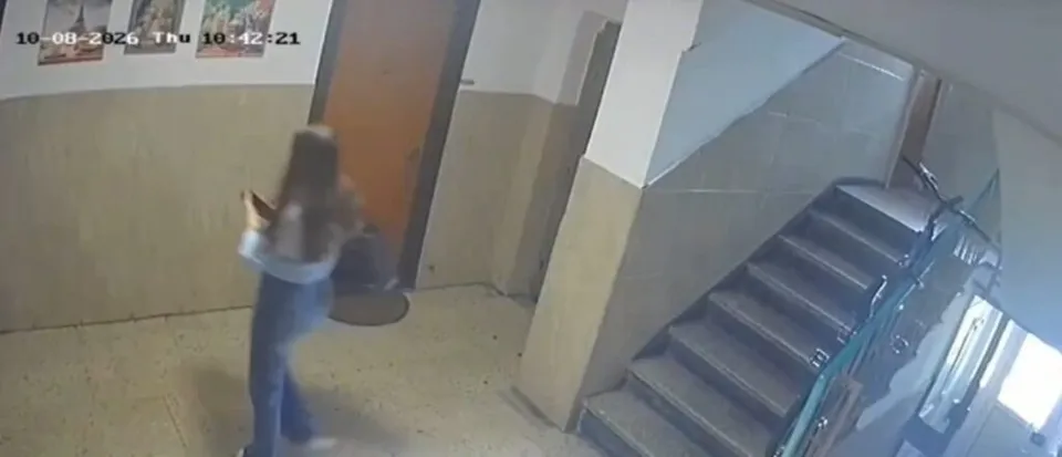 O persoană cu părul lung, purtând bluză deschisă la culoare și pantaloni albaștri, merge pe un hol lângă o scară interioară.