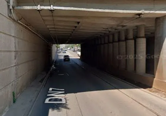 O mașină neagră circulă pe un drum asfaltat sub o structură de beton cu coloane laterale și marcaje rutiere pe suprafață.