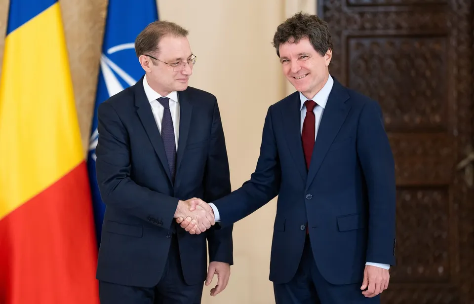 Doi bărbați în costume oficiale stau în picioare și se strâng pe mână în fața unor steaguri.