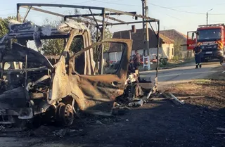 O autoutilitară arsă și carbonizată stă pe marginea unui drum lângă un camion de pompieri și câteva case.