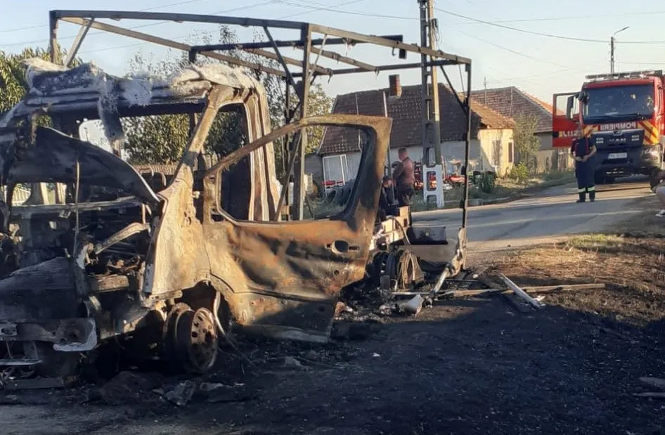O autoutilitară arsă și carbonizată stă pe marginea unui drum lângă un camion de pompieri și câteva case.