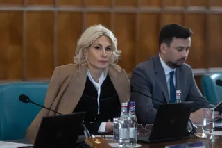 O femeie cu părul blond și o jachetă bej stă la o masă lângă un bărbat cu costum gri și cravată albastră.