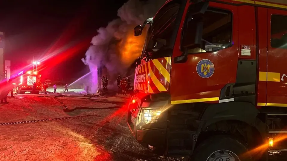 Un camion de pompieri roșu este parcat pe un drum de țară lângă coloane de fum dens sub lumini de alertă.