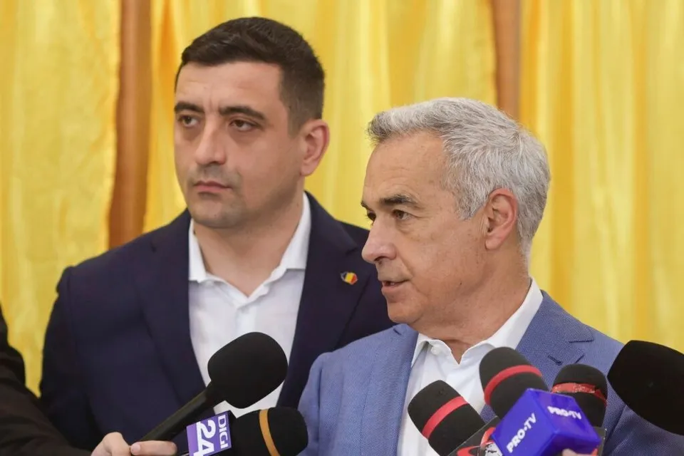 Un bărbat cu părul cărunt și sacou albastru vorbește în fața mai multor microfoane, în timp ce un alt bărbat stă în spatele lui.