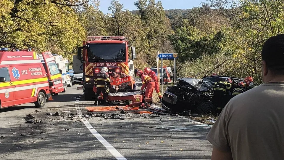 Vehicule de serviciu, inclusiv o ambulanță și un camion de pompieri, sunt parcate pe un drum lângă două mașini accidentate și persoane în uniforme de protecție.